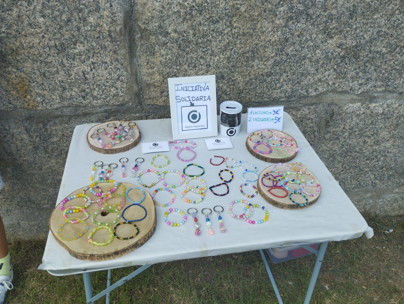 Stand solidario con pulseras y llaveros de colores sobre rodajas de madera, con el cartel de Iniciativa Solidaria de Objetivo Diagnóstico.